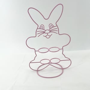 May include: A pink metal bunny rabbit egg holder with two rows of circular openings for holding eggs. The bunny has a cute cartoon face with whiskers.