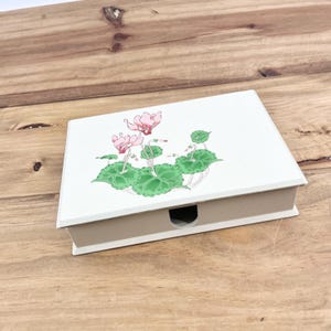 May include: A white rectangular jewelry box with a floral design on the lid. The design features pink flowers and green leaves. The box has a small, curved opening on the front and is set on a wooden surface.
