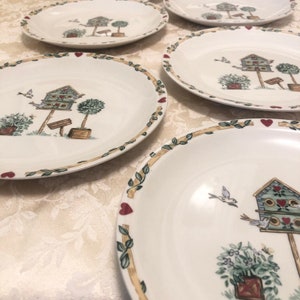 May include: Set of white ceramic plates with a birdhouse and garden design. Each plate features a birdhouse, potted plants, and a border of leaves, hearts, and vines. The plates are oval-shaped.