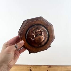 May include: An octagonal wooden plaque featuring a circular copper relief in the center. The copper inlay depicts a detailed tea kettle with a handle and spout. The wood frame has a natural brown finish with visible grain patterns throughout.