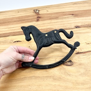 May include: Black cast iron rocking horse wall hook. The decorative hook is in the shape of a rocking horse with a saddle and curved rockers. The hook is designed to be mounted on a wall and measures approximately 7 inches long.