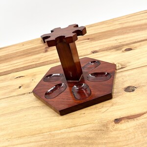 May include: A dark brown wooden pipe stand with a hexagonal base and a central support. The stand has six oval-shaped indentations for pipes and a top piece with a unique, decorative shape. The wood grain is visible.
