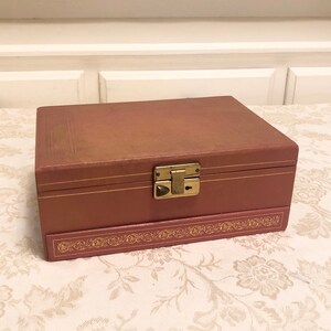 May include: A vintage pink jewelry box with a gold latch and decorative trim. The box is closed and sitting on a floral patterned surface.