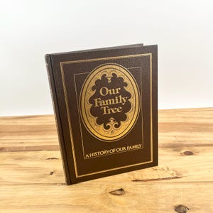 May include: A dark brown hardcover book titled Our Family Tree, A History of Our Family. The cover features a textured finish with gold foil lettering and a decorative oval border design centered on the front, standing upright on a surface.