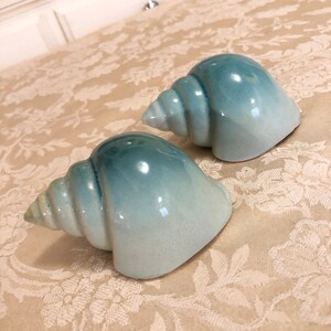 May include: Two ceramic seashells in a light blue and white ombre glaze. The shells are in a spiral shape and have a glossy finish.