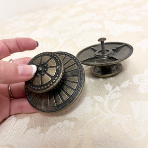 May include: Two antique brass cabinet knobs with a decorative design. The top knob has a circular design with a flower in the center. The bottom knob has a circular design with a sunburst pattern.