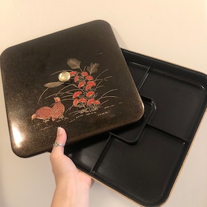 May include: A black lacquered box with a gold and red floral design and a bird on the lid. The box has a removable lid and a compartmentalized interior.