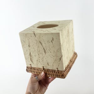 May include: A square tissue box cover featuring a cream-colored, textured stone-like top section with a circular opening and a woven brown rattan base. The two materials are connected by thin, light-colored cord loops along the bottom edge.