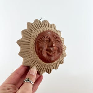 May include: A terracotta sun face wall hanging with a smiling expression. The sun has a brown face and a tan, ridged outer ring. A small wire loop is attached for hanging. The sun is held in a hand.