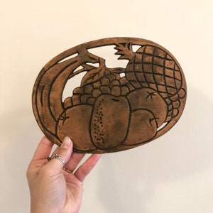 May include: A brown metal trivet with a design of fruit, including a pineapple, grapes, and pears.
