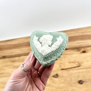 May include: A heart-shaped trinket box in a pale green colour with a white cameo design. The cameo features a woman's profile and floral accents. The box has a decorative border and is held in a hand.