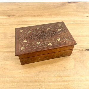 May include: A wooden box with a hinged lid, intricately carved with a floral design and inlaid with brass hearts. The box has a dark brown finish and is decorated with gold accents.