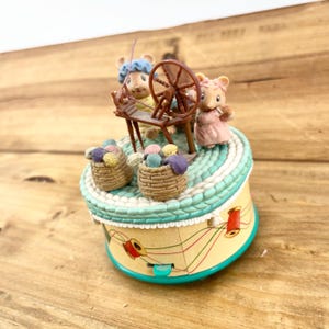 May include: A decorative music box featuring two mouse figurines, one operating a spinning wheel and the other holding yarn. The box has a light blue and white trim, two baskets of yarn, and a yellow base with thread and spool details.