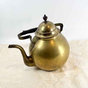 May include: A vintage brass teapot with a wooden handle and a domed lid. The teapot has a round body and a long spout.