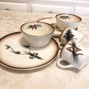 May include: A set of white ceramic dishes with a brown and green pine cone design. The set includes a large plate, a smaller plate, a bowl, and two mugs.