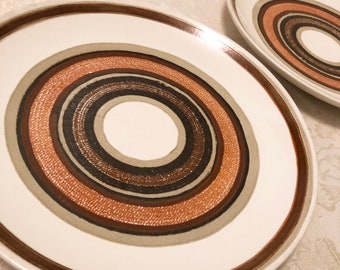 May include: Two white ceramic plates with concentric circles in shades of brown, black, and gray. The plates have a brown rim and a white center. The plates are on a white surface.