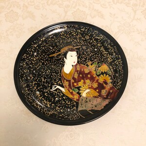 May include: A black round plate with a gold and black speckled background. A geisha in a red and gold kimono is painted on the plate.