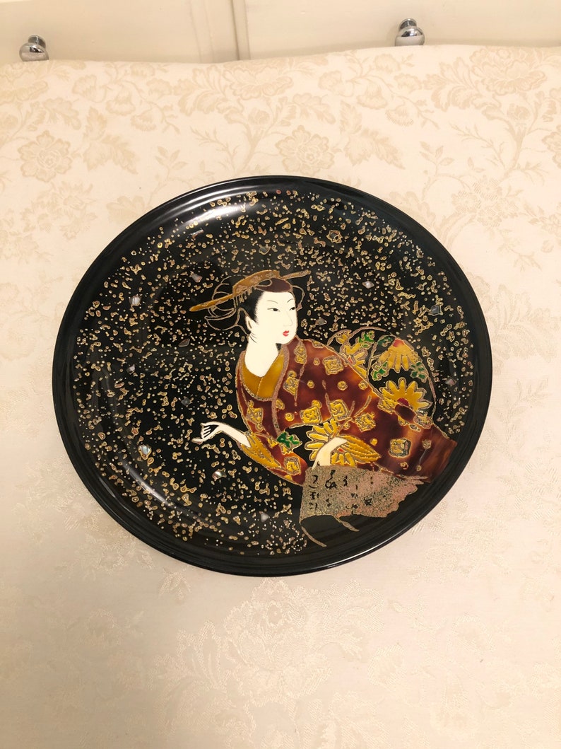 May include: A black round plate with a gold and black speckled background. A geisha in a red and gold kimono is painted on the plate.