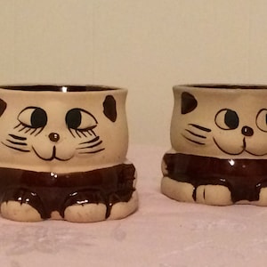 May include: Two ceramic mugs shaped like cats with brown and white paint. The mugs have brown eyes and a brown nose. The mugs are sitting on a white surface.