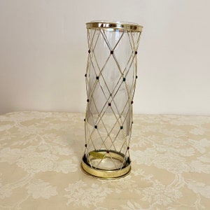May include: A clear glass vase with a gold metal cage design. The cage has a diamond pattern and is decorated with red, blue, and green jewels.