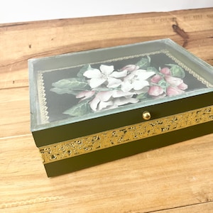 May include: A rectangular dark green box with a glass lid featuring a floral print of white and pink apple blossoms on a black background. The box is decorated with a wide, ornate gold metallic trim around the sides and a small gold knob.
