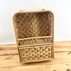 May include: A natural woven wall shelf with two compartments. The top compartment is open and the bottom compartment has a door. The shelf is made of natural woven materials and has a light brown color.