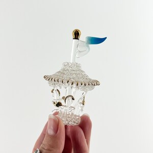 May include: A miniature glass carousel with a gold-colored top and accents. The carousel features small horses and a blue and white flag. The carousel is held in a person's hand.