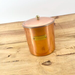 May include: A cylindrical copper canister with a matching lid and a small round brass knob on top. A rectangular brass plaque is attached to the front of the canister with the word SUGAR embossed in capital letters on the metal surface.