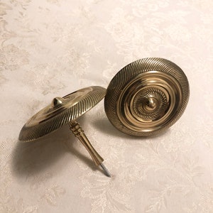 May include: Two gold-toned metal curtain tiebacks with a decorative, circular design. Each tieback has a screw-on attachment.