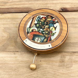 May include: A wooden music box with a round, brown lid featuring a colorful illustration of angels singing around a manger. The lid is inscribed with "Christmas 1976".