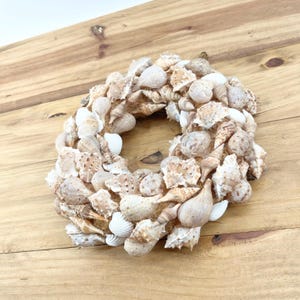 May include: A decorative wreath composed of various seashells in shades of white, beige, and brown. The shells are arranged in a circular shape, creating a natural, coastal aesthetic. The wreath is set against a wooden background.