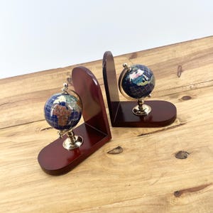 May include: Two decorative globe bookends. Each bookend features a small globe with a blue ocean and gold stand, set on a dark wood base. The bookends are made of dark wood and are designed to hold books upright.