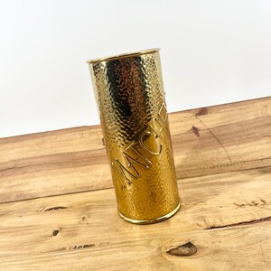 May include: A gold-colored match holder with a hammered texture. The word "MATCHES" is embossed on the side. The holder is cylindrical and sits on a wooden surface.