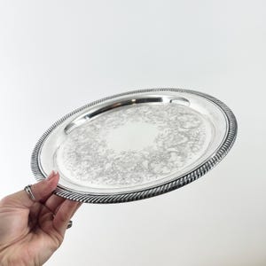 May include: A round silver-toned serving tray featuring a polished finish and an intricate engraved floral pattern across the center. The edge of the tray is defined by a raised, textured rope-style border that encircles the entire circumference.