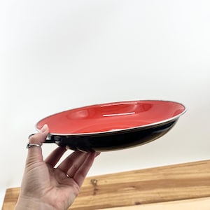 May include: A red and black enamelware plate with a silver rim. The plate is held up against a white background. The plate has a handle on the side.