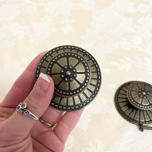 May include: Two antique gold-toned metal cabinet knobs with a circular design. The knobs have a raised center with a circular pattern and a decorative outer ring.