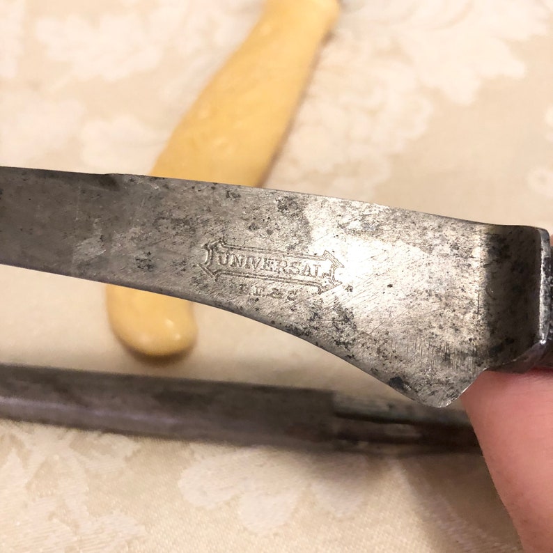 May include: Close-up of a vintage knife blade with a worn metal finish. The blade is marked with the word "UNIVERSAL" in a decorative font.