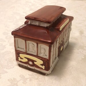 K&ouml;nnte beinhalten: Ein braunes Keramik-Sparschwein in Form einer San Francisco-Stra&szlig;enbahn. Die Bahn hat Fenster mit bemalten Figuren im Inneren und einen gelben Streifen mit den Buchstaben "S.F."