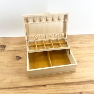 May include: An open, off-white jewelry box with a hinged lid. The interior features a light tan fabric lining and a pull-out tray with small compartments. The base has a decorative trim. The box is on a wooden surface.