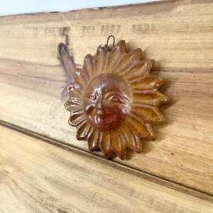 May include: A circular sun-shaped wall plaque with a warm brown and orange glazed finish. The sun features a smiling face with closed eyes and a nose, surrounded by pointed rays. A small metal wire loop is attached to the top for hanging.