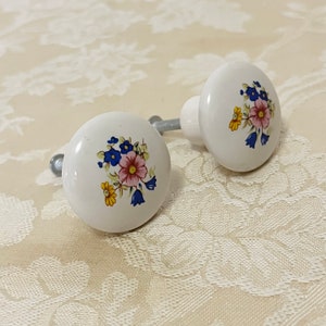 May include: Two white ceramic cabinet knobs with floral designs. The knobs feature a bouquet of pink, yellow, and blue flowers on a white background.