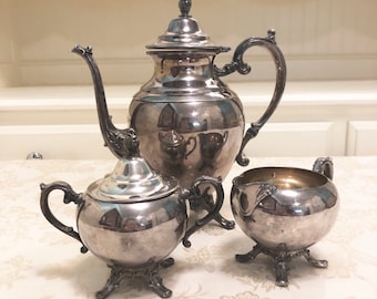 May include: A silver tea set with a teapot, creamer, and sugar bowl. The set has a traditional design with ornate details and a polished finish.