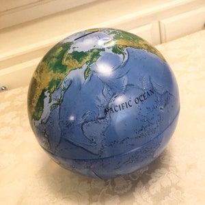 May include: A blue globe piggy bank with a green and brown map of the world. The globe has a slot on the top for coins and the words "Pacific Ocean" are printed on the side.