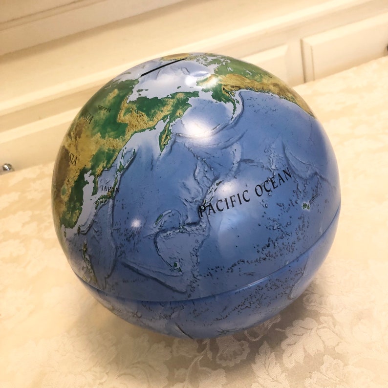 May include: A blue globe piggy bank with a green and brown map of the world. The globe has a slot on the top for coins and the words "Pacific Ocean" are printed on the side.