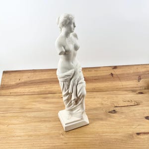 May include: A white ceramic statue of a classical female figure, missing arms, draped in fabric. The statue stands on a square base and is set against a wooden surface and white background. The figure is a representation of classical art.