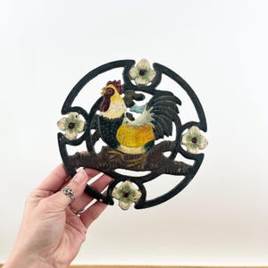 May include: A cast iron trivet featuring a colorful rooster perched on a branch, surrounded by a black circular frame with floral accents. The rooster has yellow, black, and white plumage with a red comb. The trivet is designed to protect surfaces from hot cookware.
