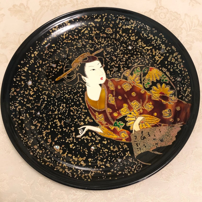 May include: A black lacquer plate with gold and white accents features a painted image of a woman in a red and gold kimono. The plate has a speckled background and the woman is surrounded by gold and white dots.