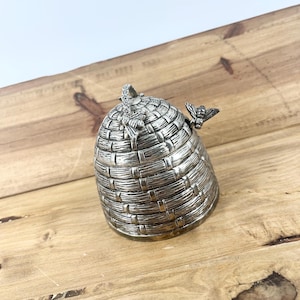 May include: A silver-toned metal honey pot shaped like a traditional skep beehive. The exterior features a textured, woven basket pattern with a small bee figurine attached to the top handle and another bee figurine resting on the side rim.