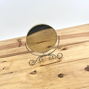 May include: A small, round vanity mirror with a gold-colored metal frame and base. The mirror is supported by a decorative, scrolled stand. The mirror is set against a light-colored wooden surface.