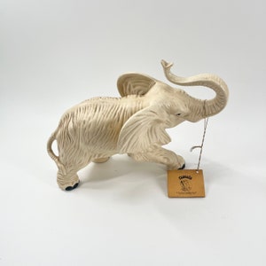 May include: White ceramic elephant figurine with its trunk raised in the air. The figurine is standing on a white surface and has a small brown tag with the word "Rascal's" printed on it.